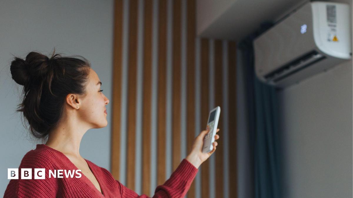 A woman with brown hair, wearing a red cardigan, is turning on a wall-mounted air-conditioning unit with a remote.