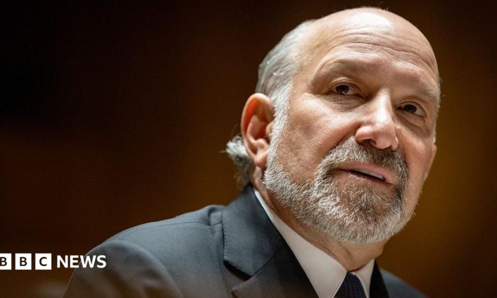 Close-up portrait of Howard Lutnick with a short grey beard and receding hairline, wearing a dark suit jacket, white shirt, and dark tie. The person is turned slightly to the side and looking toward the camera. The background is softly blurred with warm brown and amber tones, creating a shallow depth-of-field effect.