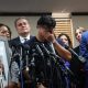 A man holds his hands over his eyes as he stands before a bank of microphones and a man holding up pictures of Lorenzo Salgado Araujo