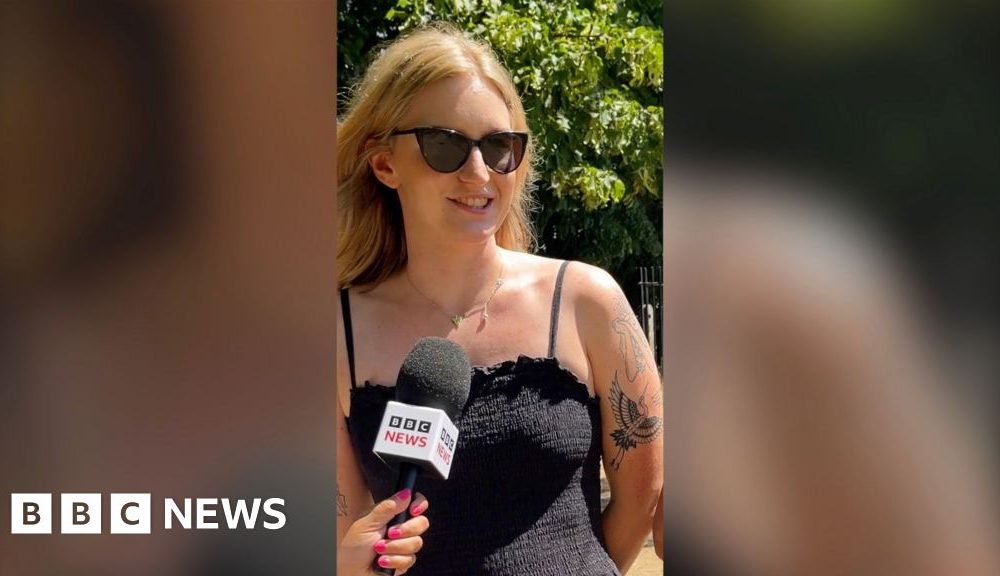 A woman speaking into a mic. She has blonde hair, sunglasses, a black strappy dress and tattoos.