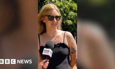 A woman speaking into a mic. She has blonde hair, sunglasses, a black strappy dress and tattoos.