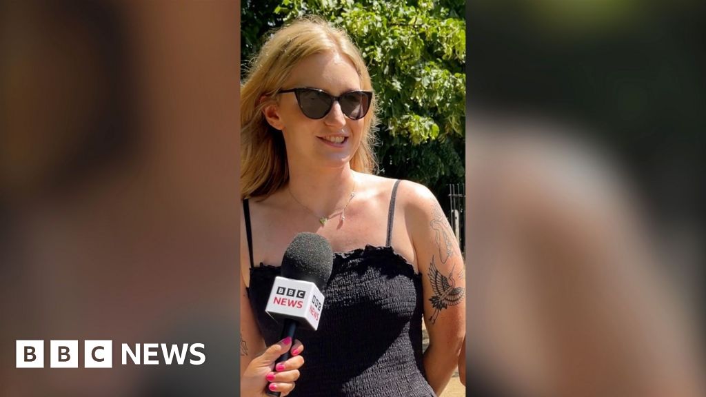 A woman speaking into a mic. She has blonde hair, sunglasses, a black strappy dress and tattoos.