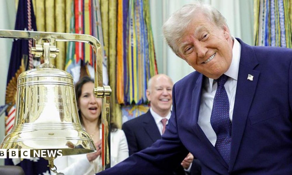 President Donald Trump ringing the opening bell in the Oval Office on Monday. The US president is smiling at the cameras as he rings the big gold bell. He is wearing a dark blue suite, with a dark blue spotted tie and a white shirt.