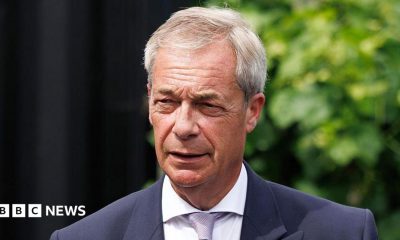 Catherine Turnbull is smiling at the camera. She is wearing a pale blue t-shirt and dark framed glasses. Catherine has short light brown hair. She has some bushes behind her with are slightly out of focus.