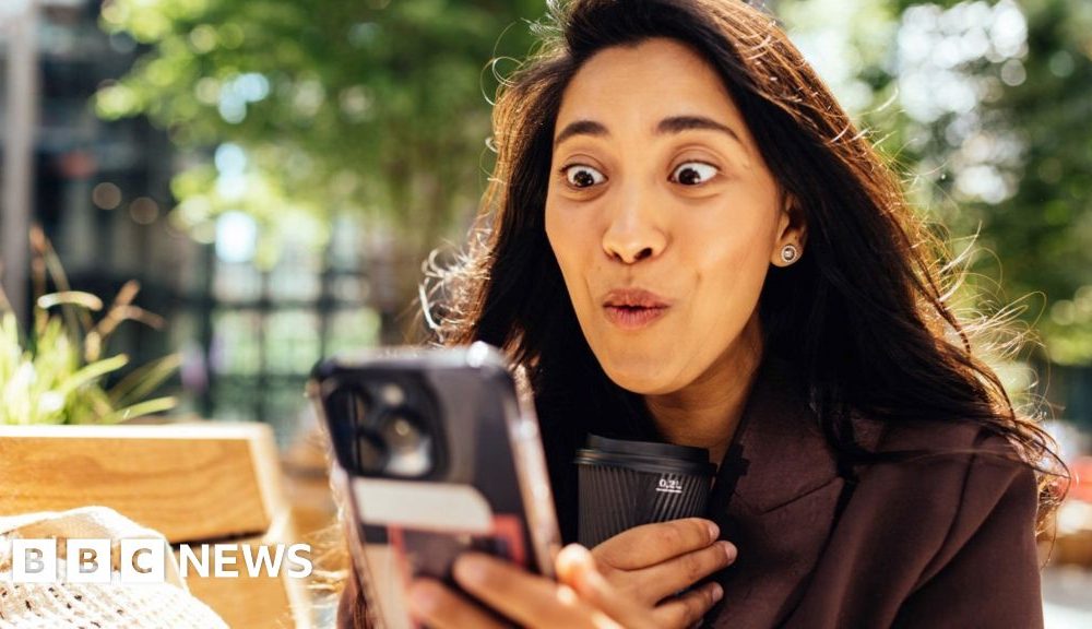 woman with long dark hair looks at her phone, with an expression of joy on her face.