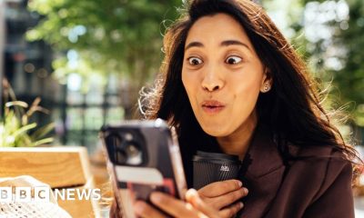 woman with long dark hair looks at her phone, with an expression of joy on her face.