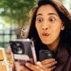 woman with long dark hair looks at her phone, with an expression of joy on her face.