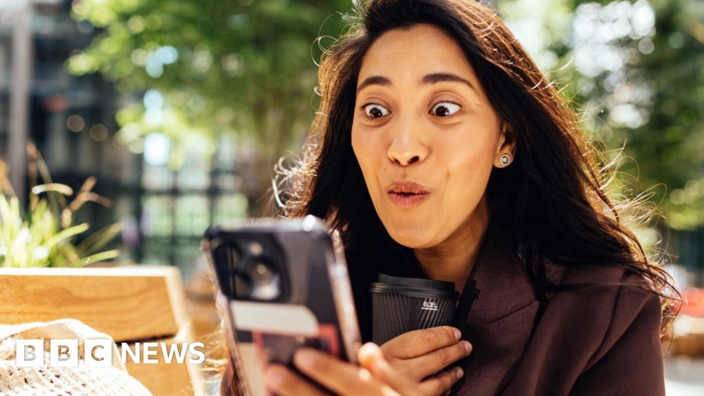 woman with long dark hair looks at her phone, with an expression of joy on her face.
