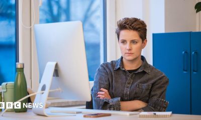 A young woman with short hair wearing a grey denim shirt working in an office, sitting with arms crossed at the desk in front on computer.