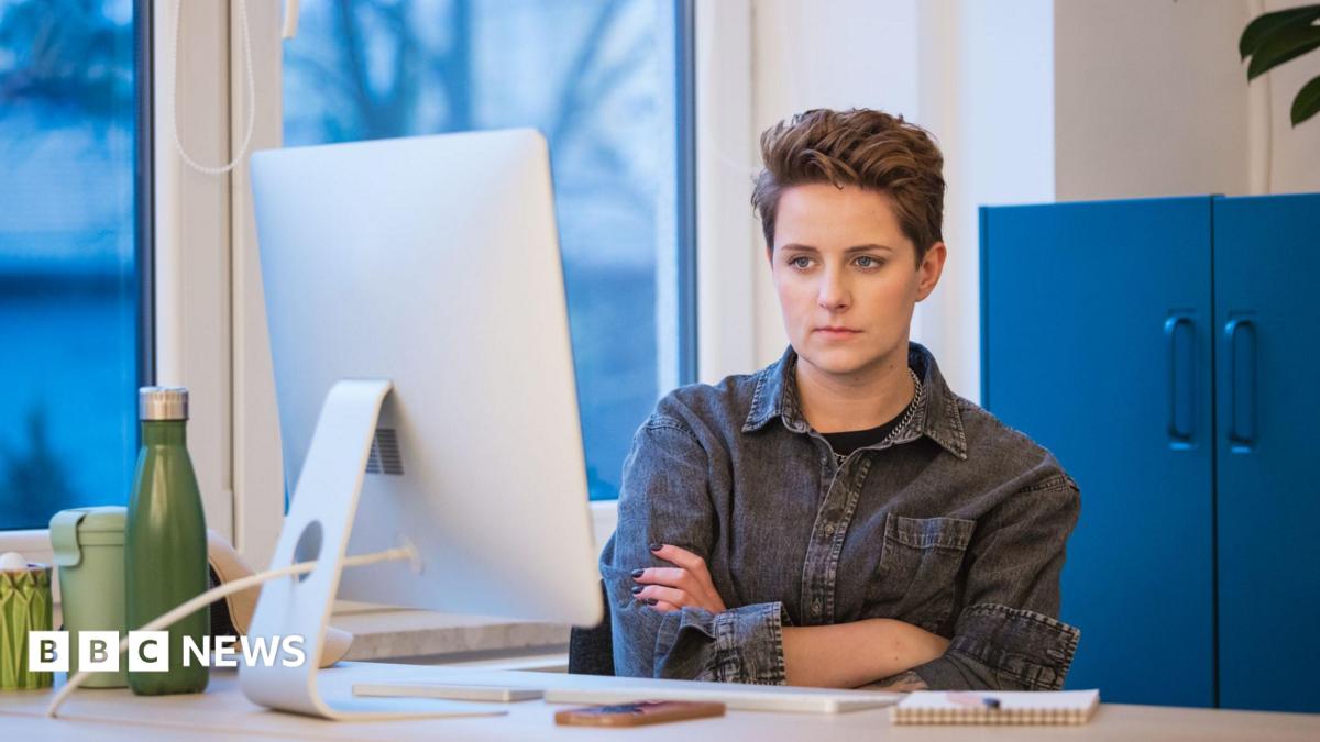 A young woman with short hair wearing a grey denim shirt working in an office, sitting with arms crossed at the desk in front on computer.