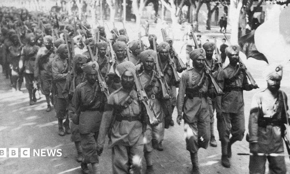 A black and white image of a large group of Indian troops, wearing soldiers' uniforms, turbans, and holding guns, walk along a road