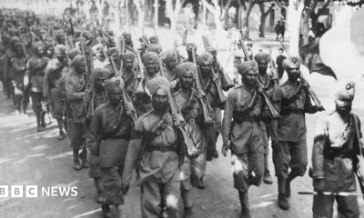 A black and white image of a large group of Indian troops, wearing soldiers' uniforms, turbans, and holding guns, walk along a road