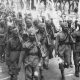 A black and white image of a large group of Indian troops, wearing soldiers' uniforms, turbans, and holding guns, walk along a road