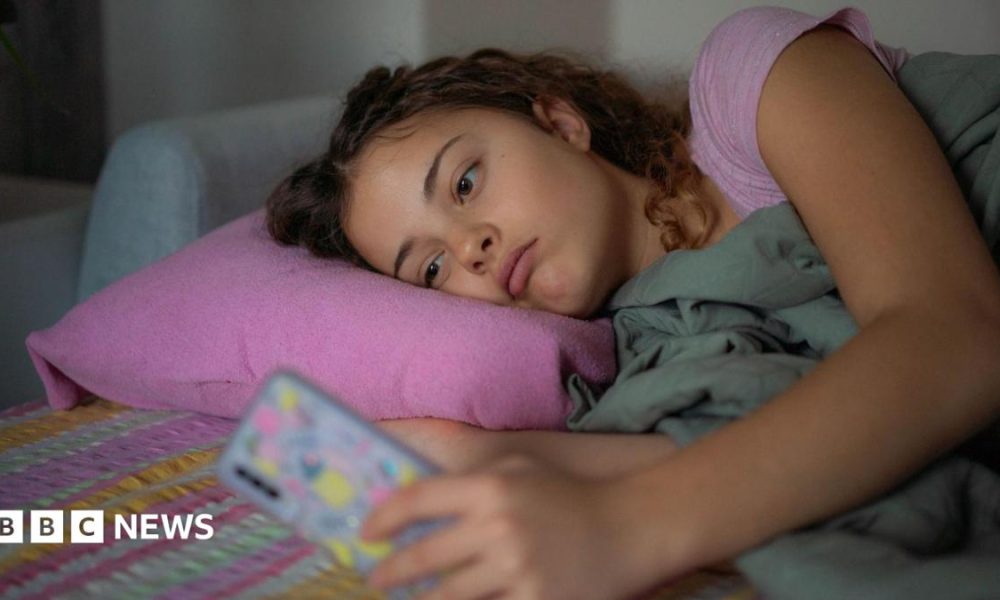 A stock image shows a young woman lying down with a glum expression as she scrolls on her smartphone