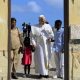 Pope Leo holds hands with a girl wearing a red dress and a man in blue shirt and white trousers. A women in a beige outfit stands alongside them at a doorway. Behind them are blue skies and men dressed in black