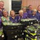 A large group of people, in standing behind a large selection of boxes of salad. They are all wearing purple T-shirts. They are in a room with items all behind them and are all smiling and looking at the camera.