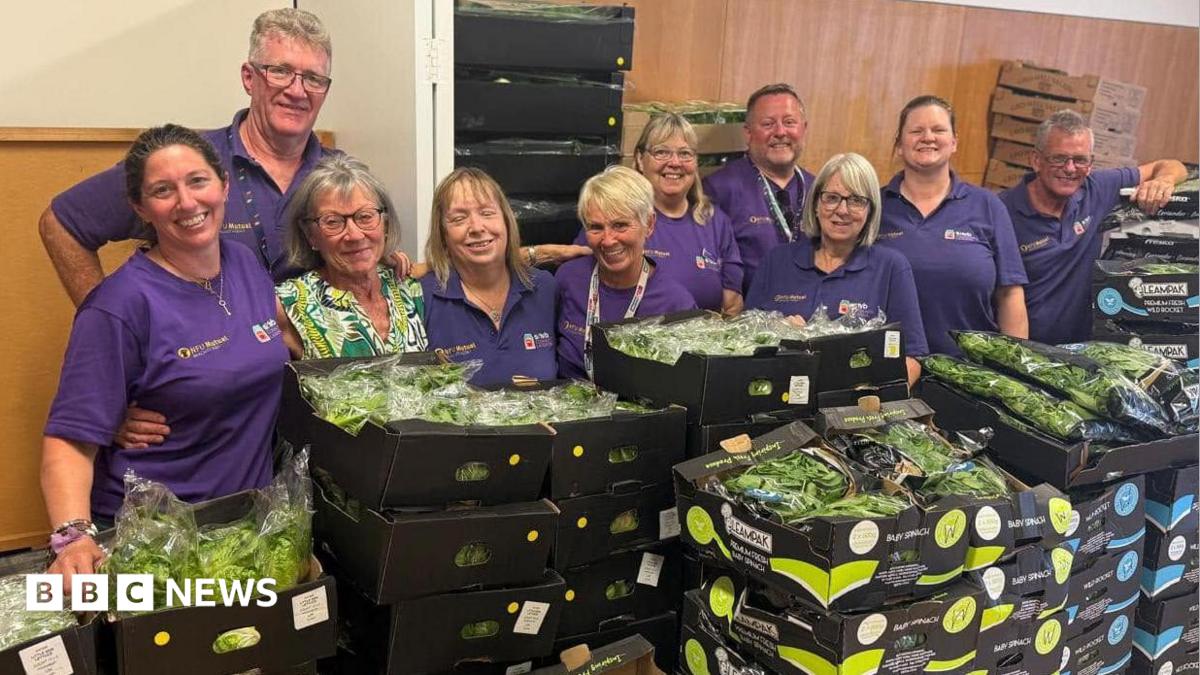 A large group of people, in standing behind a large selection of boxes of salad. They are all wearing purple T-shirts. They are in a room with items all behind them and are all smiling and looking at the camera.
