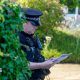 A police officer looks down at a sheet of paper while wearing uniform next to a bush and police van outside Widdecombe's home on Friday.