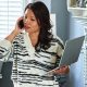 A stock image of a woman in her home talking on the phone in front of a fireplace and holding a laptop looking frustrated