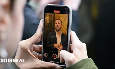 Two hands holding a phone with Zack Polanski on the screen