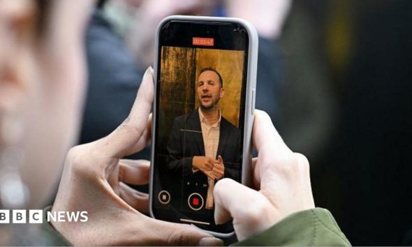 Two hands holding a phone with Zack Polanski on the screen