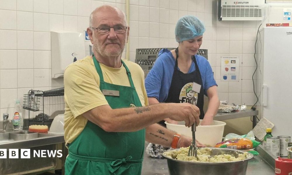 Ian White with glasses, a yellow t-shirt and green apron, preparing mashed potatoes in a large bowl while a woman with a blue hairnet, blue t-shirt and blue apron works nearby in a kitchen.