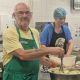 Ian White with glasses, a yellow t-shirt and green apron, preparing mashed potatoes in a large bowl while a woman with a blue hairnet, blue t-shirt and blue apron works nearby in a kitchen.
