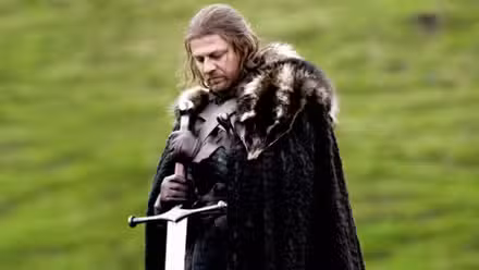 Sean Bean as Ned Stark standing in a green field and holding a sword in Game of Thrones.