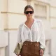 PARIS, FRANCE - MAY 26: Diane Batoukina wears long straight brown hair with a center part pulled into a low bun, black oversized square sunglasses with dark lenses by KALEOS, an ivory white sheer blouse shirt with an open collar, lace panel detailing, floral embroidery and long sleeves by Gerard Darel, high-waisted brown suede leather flared pants with front patch pockets by Gerard Darel, a beige leather clutch bag, during a street style fashion photo session, on May 26, 2026 in Paris, France. (Photo by Edward Berthelot/Getty Images)