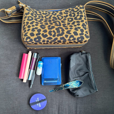 image of what's in Kat's bag on July 1, 2026: black pouch, blue wallet, blue hair clip, purple mirror with elastic wrapped around it, 2 lip glosses, concealer, and a (very old) pill case. The purse itself is leopard from Rothy's.