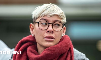 A woman with short blonde hair and glasses, wearing a grey court and red scarf