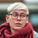 A woman with short blonde hair and glasses, wearing a grey court and red scarf