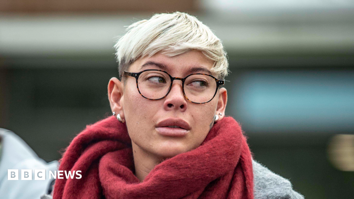 A woman with short blonde hair and glasses, wearing a grey court and red scarf