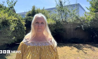 A woman with long blonde hair stands in her garden. Behind her, beyond her garden fence, is a large data centre which looks like a tall warehouse