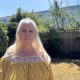 A woman with long blonde hair stands in her garden. Behind her, beyond her garden fence, is a large data centre which looks like a tall warehouse