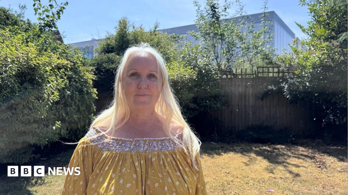 A woman with long blonde hair stands in her garden. Behind her, beyond her garden fence, is a large data centre which looks like a tall warehouse