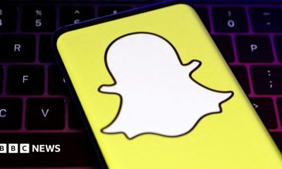 A closeup of Snapchat's logo, a white cartoon ghost with a bright yellow background, held over a laptop conputer keyboard.