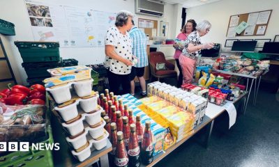 Four volunteers - three women and a man - volunteering at a foodbank. They are organising the food and drink products - which include bottles of Ribena, cartons of oat milk, and snap pots of spaghetti hoops - neatly onto a table.