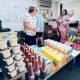 Four volunteers - three women and a man - volunteering at a foodbank. They are organising the food and drink products - which include bottles of Ribena, cartons of oat milk, and snap pots of spaghetti hoops - neatly onto a table.