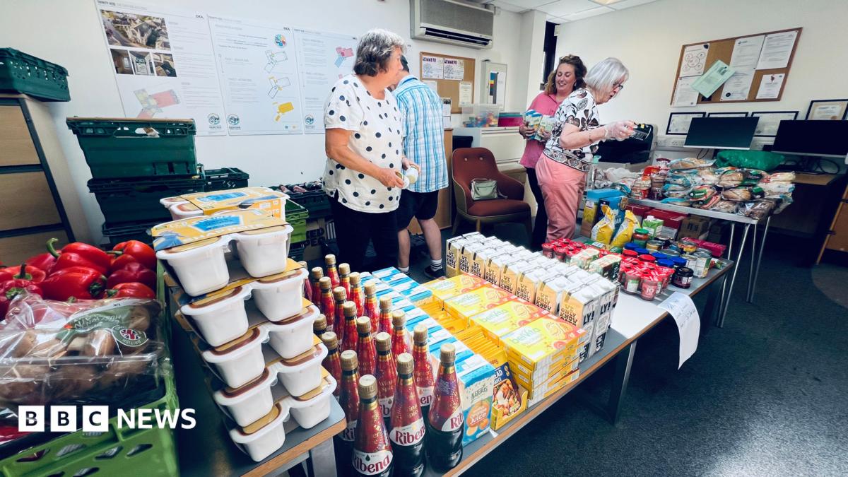 Four volunteers - three women and a man - volunteering at a foodbank. They are organising the food and drink products - which include bottles of Ribena, cartons of oat milk, and snap pots of spaghetti hoops - neatly onto a table.