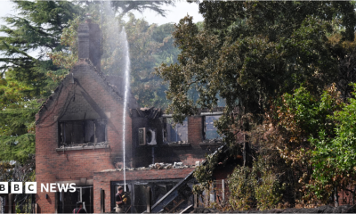 An aerial image shows several houses destroyed by fire, with their roofs gone and wreckage visible within. There is a line of dead trees next to the houses and areas of scorched earth. A fire engine is parked by one of the houses.