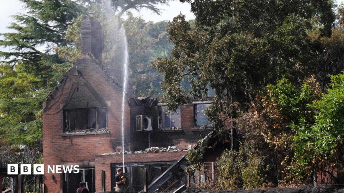 An aerial image shows several houses destroyed by fire, with their roofs gone and wreckage visible within. There is a line of dead trees next to the houses and areas of scorched earth. A fire engine is parked by one of the houses.