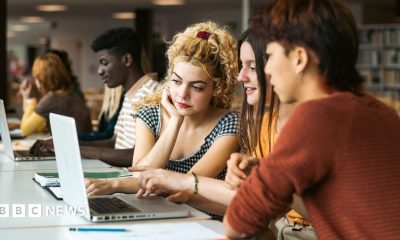 Stock photo shows three students looking at a laptop in a university library with other students and book shelves in the background.