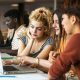 Stock photo shows three students looking at a laptop in a university library with other students and book shelves in the background.