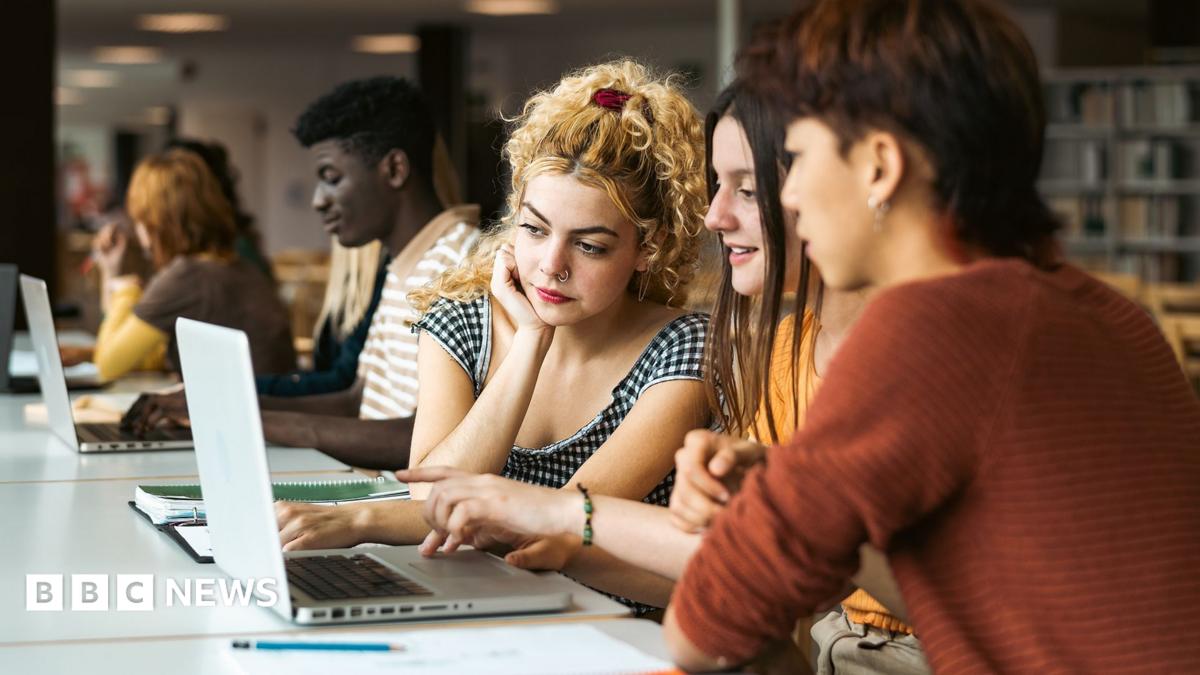 Stock photo shows three students looking at a laptop in a university library with other students and book shelves in the background.