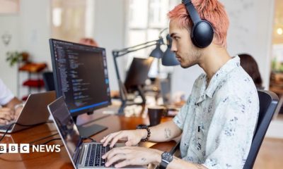 A tech worker with pink hair sits at a desk and works on a laptop