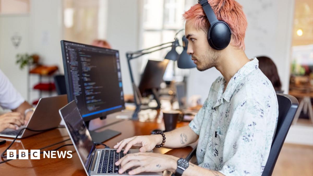 A tech worker with pink hair sits at a desk and works on a laptop