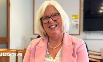 Dr Nicola Brink is wearing a pink jacket as she sits in a room and smiling directly at the camera. She has pale blonde hair and glasses.