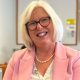 Dr Nicola Brink is wearing a pink jacket as she sits in a room and smiling directly at the camera. She has pale blonde hair and glasses.
