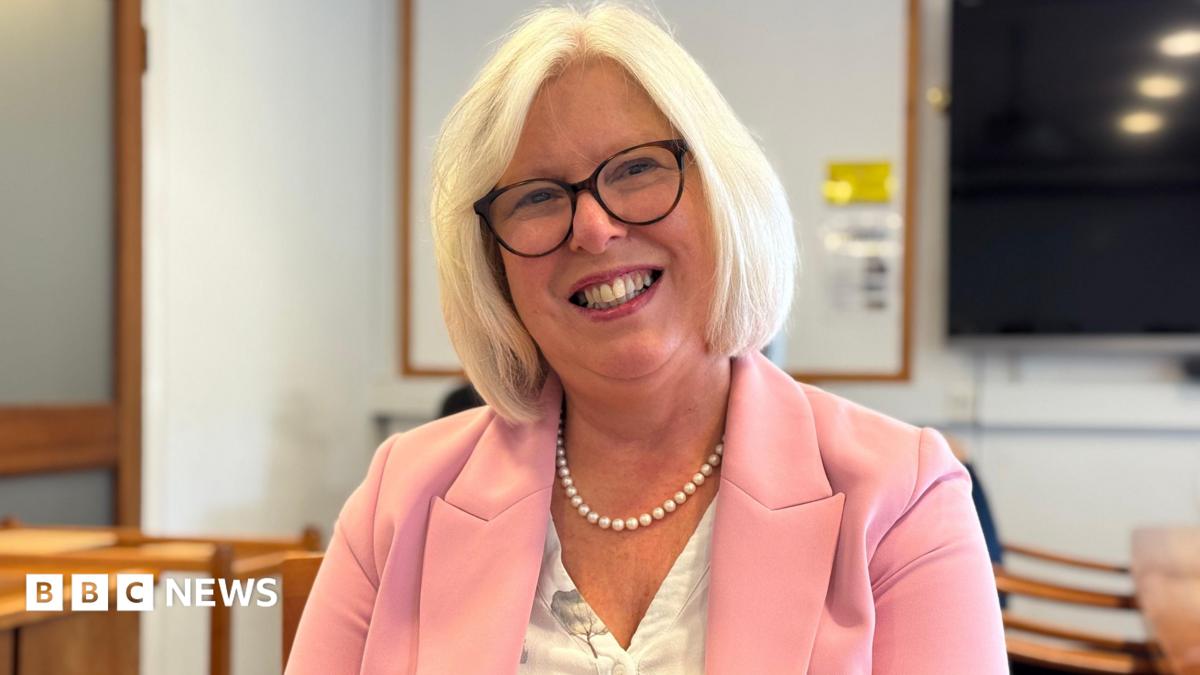 Dr Nicola Brink is wearing a pink jacket as she sits in a room and smiling directly at the camera. She has pale blonde hair and glasses.
